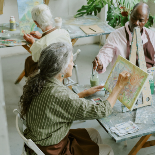 group of older adults in an art studio painting on canvas. Older-adult woman in foreground painting flowers on canvas
